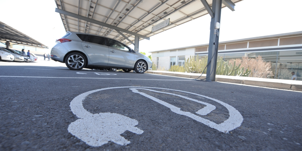 Parking del ITER con punto de recarga y coche eléctrico en segundo plano y el símbolo de punto de recarga pintado en el cemento del suelo en primer término.