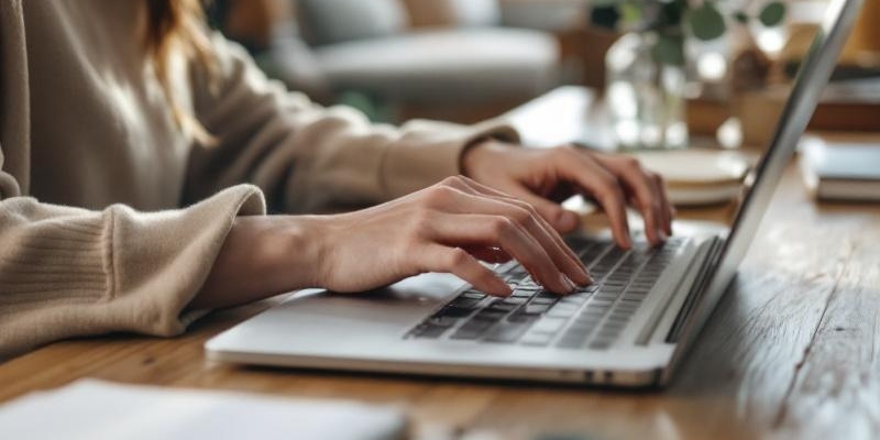 Manos femeninas tecleando en laptop plateado sobre escritorio de madera con luz natural matutina.