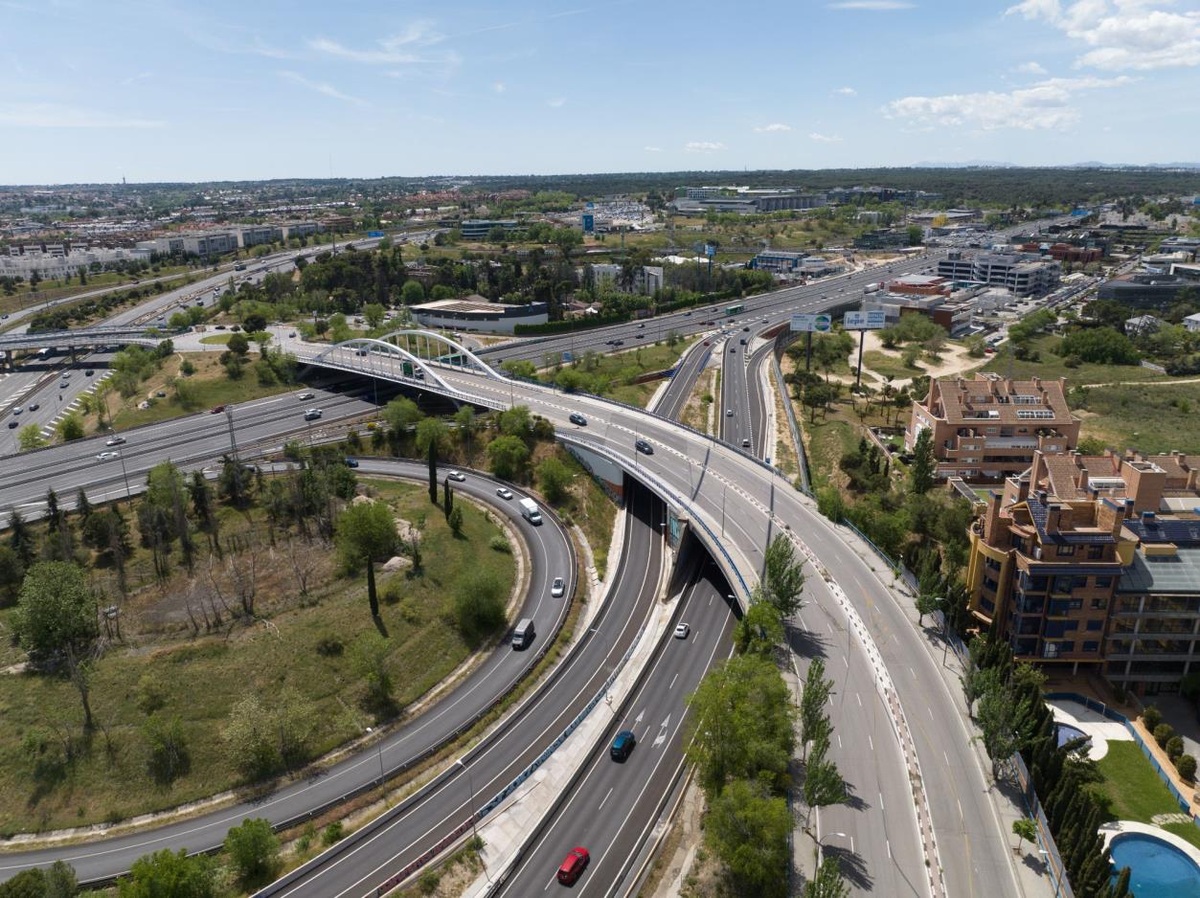 Vista aérea de un enlace viario con vehículos, puentes y ramales de acceso junto a edificios y zonas verdes.