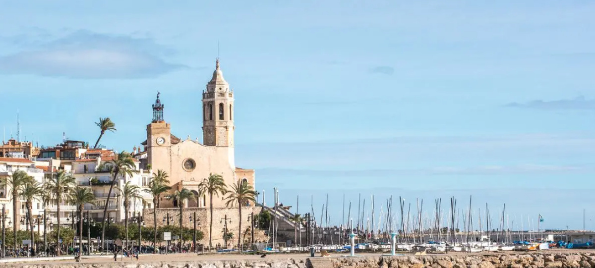Vista del frente marítimo de Sitges con la iglesia, palmeras y embarcaciones junto a la costa.