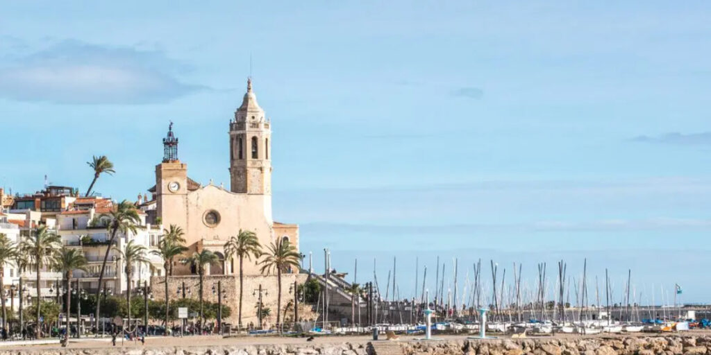 Vista del frente marítimo de Sitges con la iglesia, palmeras y embarcaciones junto a la costa.